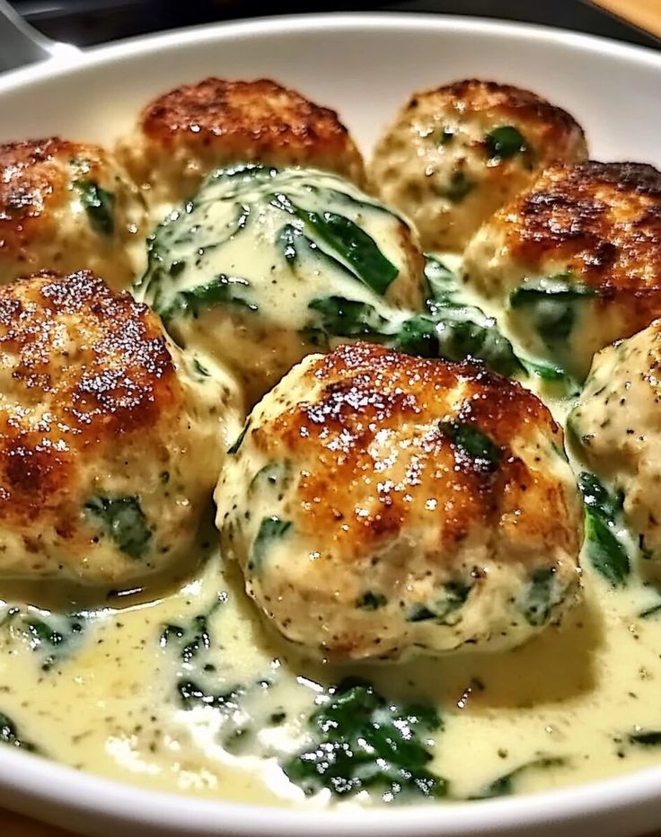 a white bowl filled with meatballs covered in cheese and spinach sauce on top of a wooden table