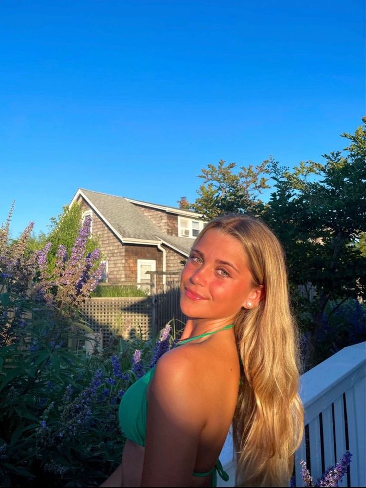 a beautiful young blond woman standing in front of a house with purple flowers on it