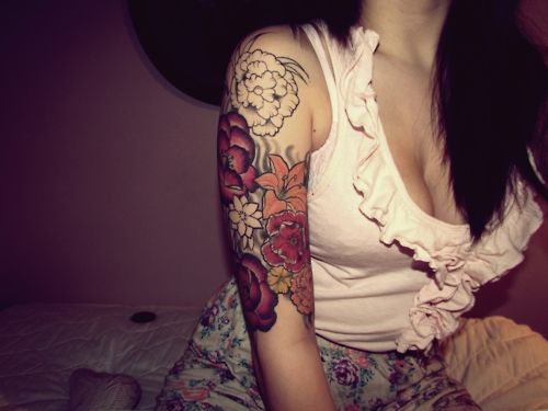 a woman with a flower tattoo on her arm, sitting on a bed and looking at the camera