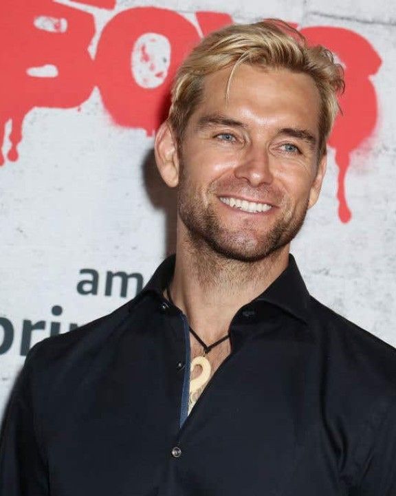 a man with blonde hair smiles at the camera while standing in front of a wall