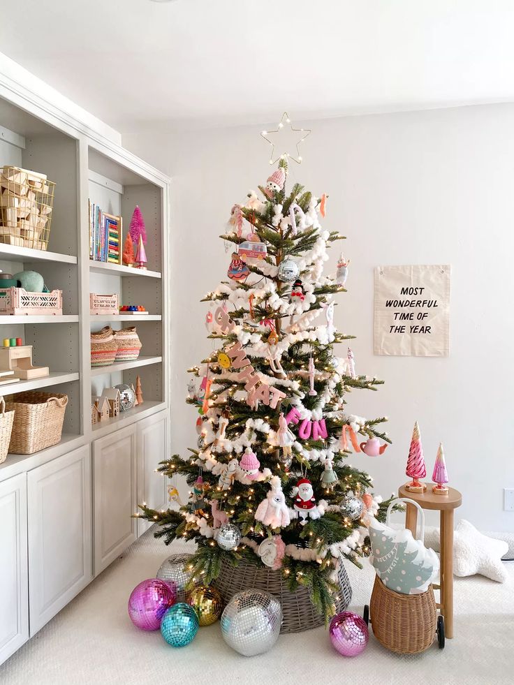 a white christmas tree with pink and blue ornaments in a living room next to bookshelves