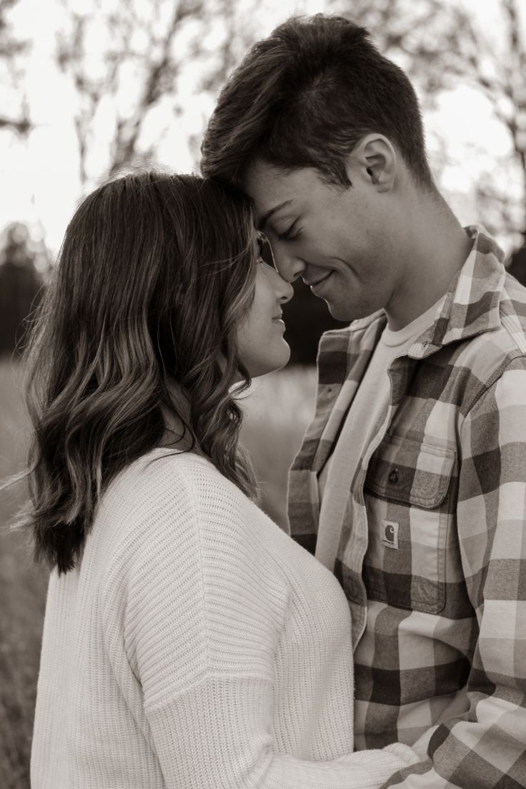 a man and woman standing next to each other in front of some trees with their faces close together