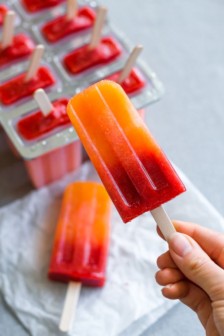 a hand holding an orange and red popsicle next to trays of ice cream