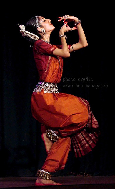 a woman in an orange and red outfit dancing with her hands on her hip,