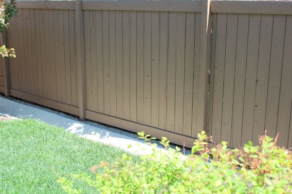 a brown fence in front of a green lawn and shrubbery on the side of a house