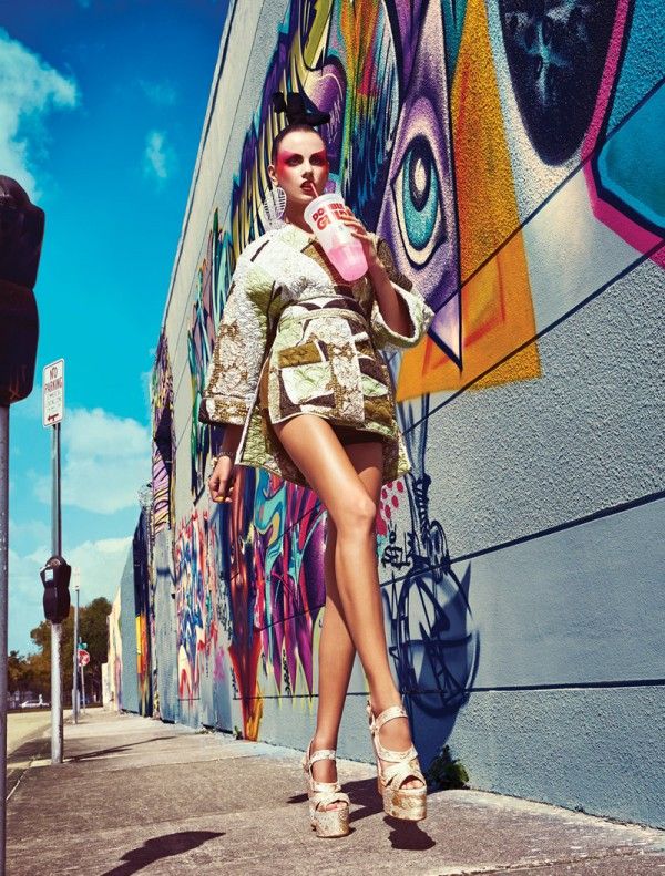 a woman standing on the side of a building with her legs crossed and drinking from a cup