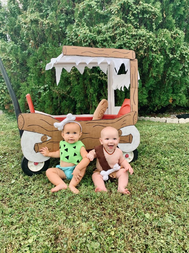 two babies sitting on the grass in front of a wooden toy truck with a cow