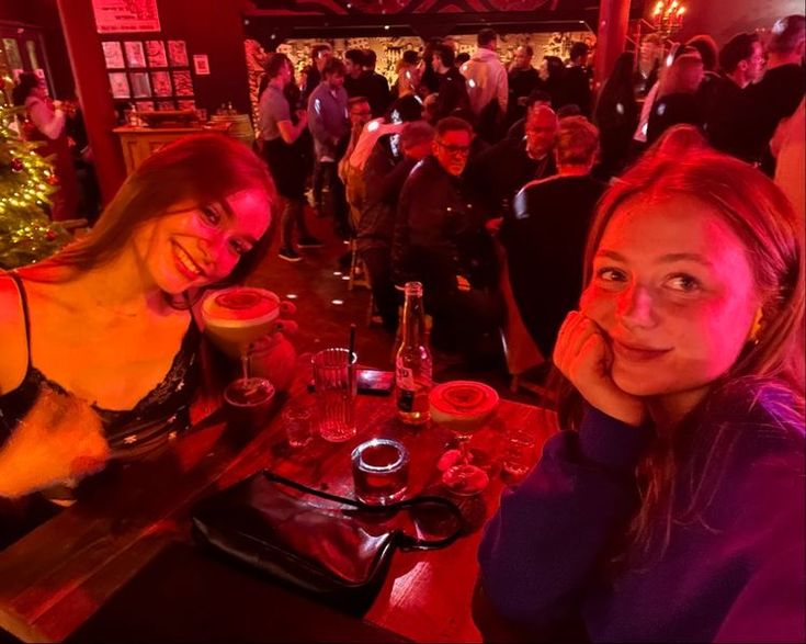 two women sitting at a table with drinks in front of them and people standing around