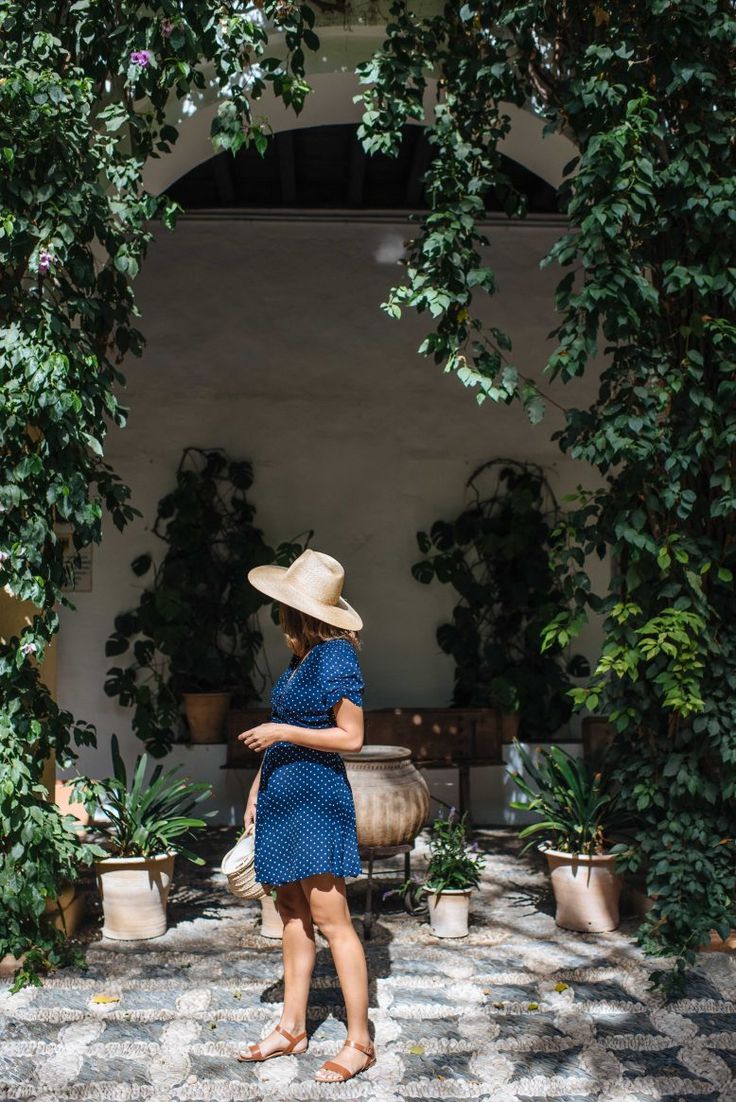 a woman in a blue dress and straw hat walking through an archway with potted plants