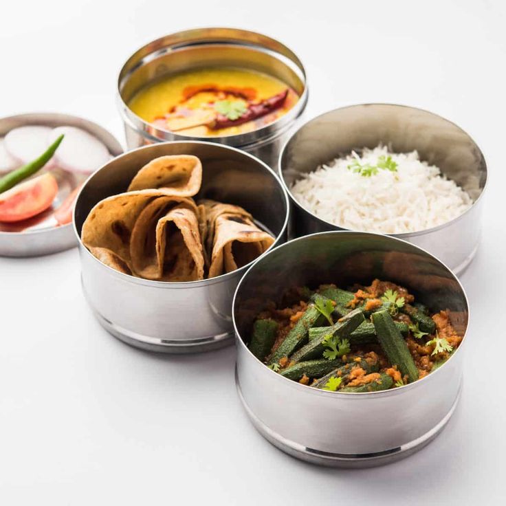 four metal containers filled with food on top of a white countertop next to rice and veggies