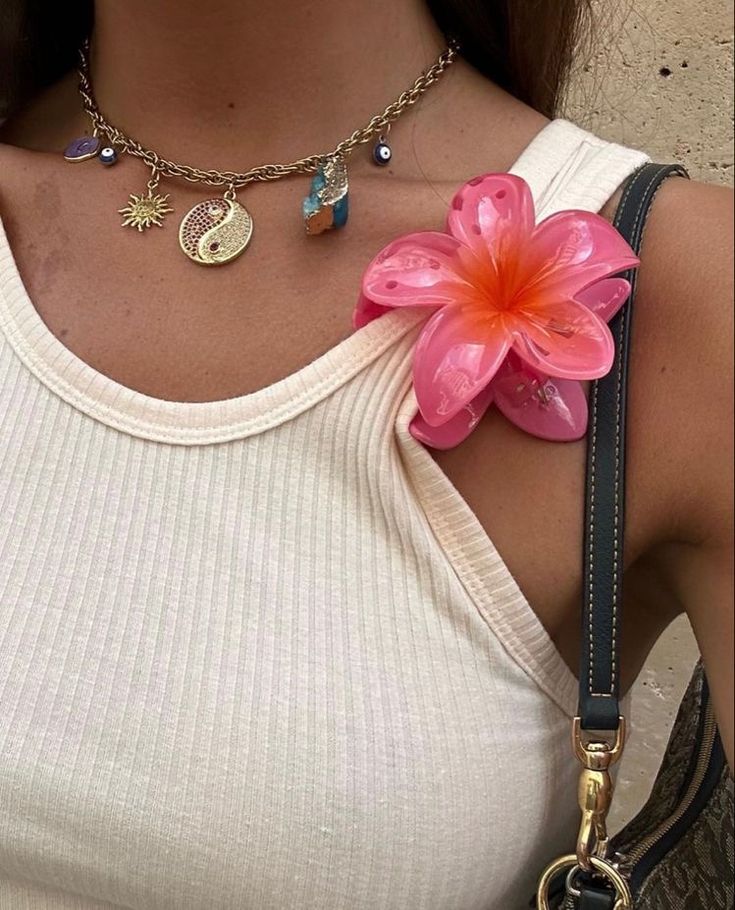 a woman wearing a white tank top holding a pink flower in her right hand and a gold chain necklace on her left shoulder