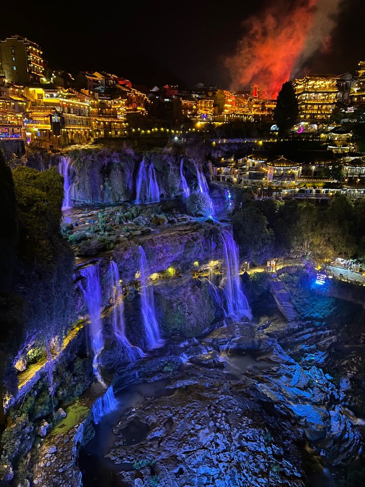 the waterfall is lit up at night with colorful lights on it's sides and buildings in the background