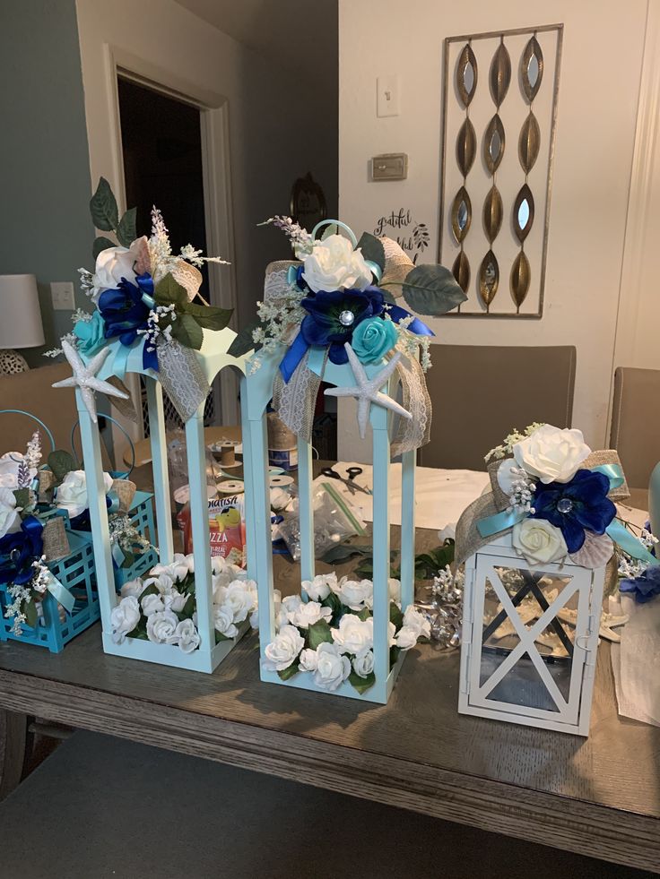 three white lanterns with blue ribbons and flowers on the top are sitting on a table