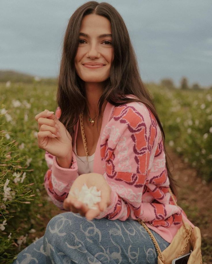 a woman is sitting in a field with flowers and holding something up to her mouth