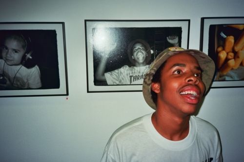 a man wearing a hat standing in front of some pictures on the wall with his mouth open