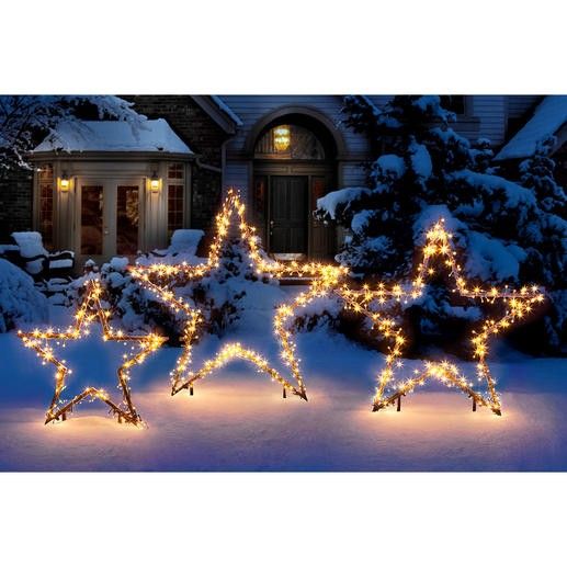 lighted stars in front of a house with snow on the ground and trees behind them