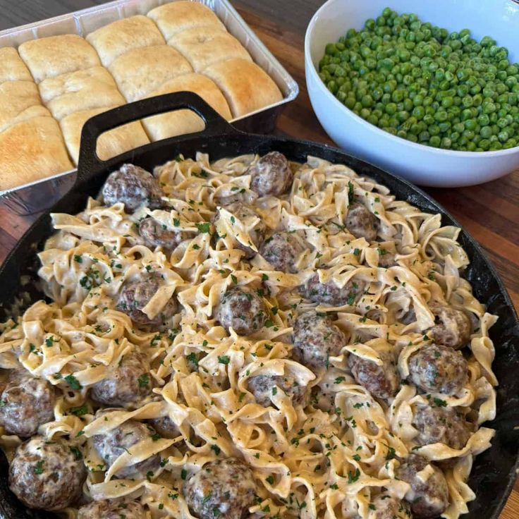 a skillet filled with pasta and meatballs next to peas, bread rolls and green peas