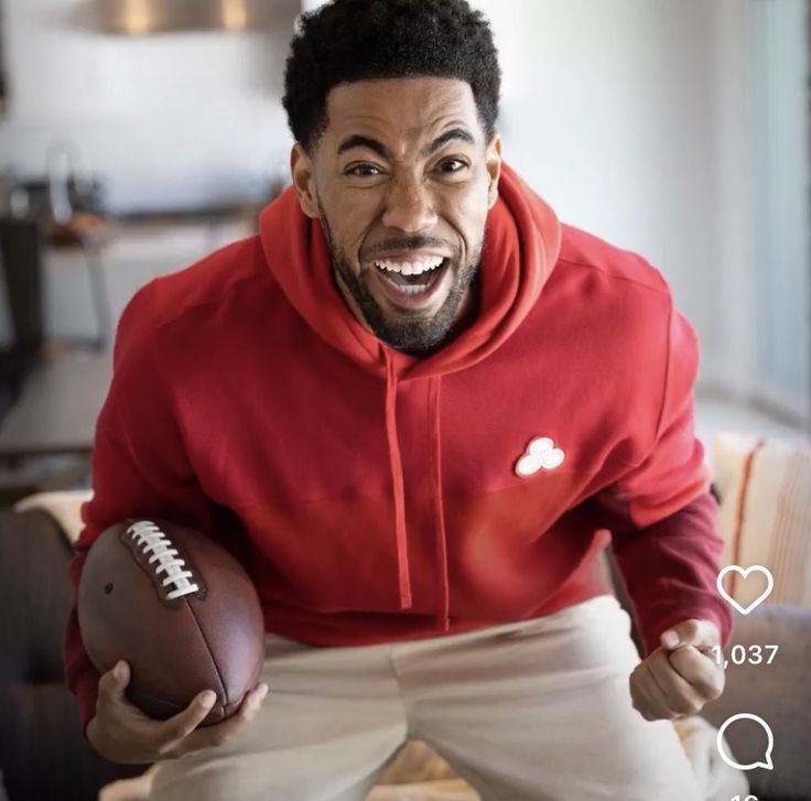 a man in a red hoodie holding a football and posing for the camera with his mouth open