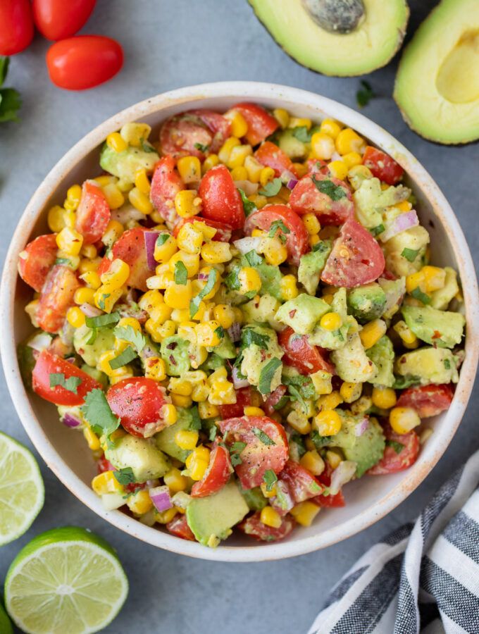 a bowl filled with corn and avocado next to sliced tomatoes, limes, and an avocado wedge