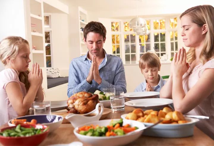 a family sitting around a dinner table with food on the table and praying in front of them