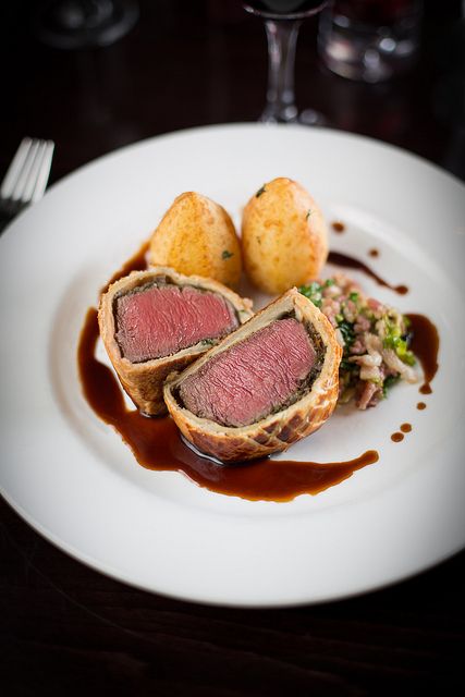 a white plate topped with meat and potatoes covered in sauce next to a glass of wine