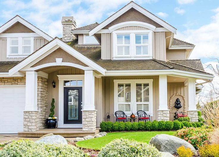 a house with white shutters and stonework on the front porch, surrounded by landscaping