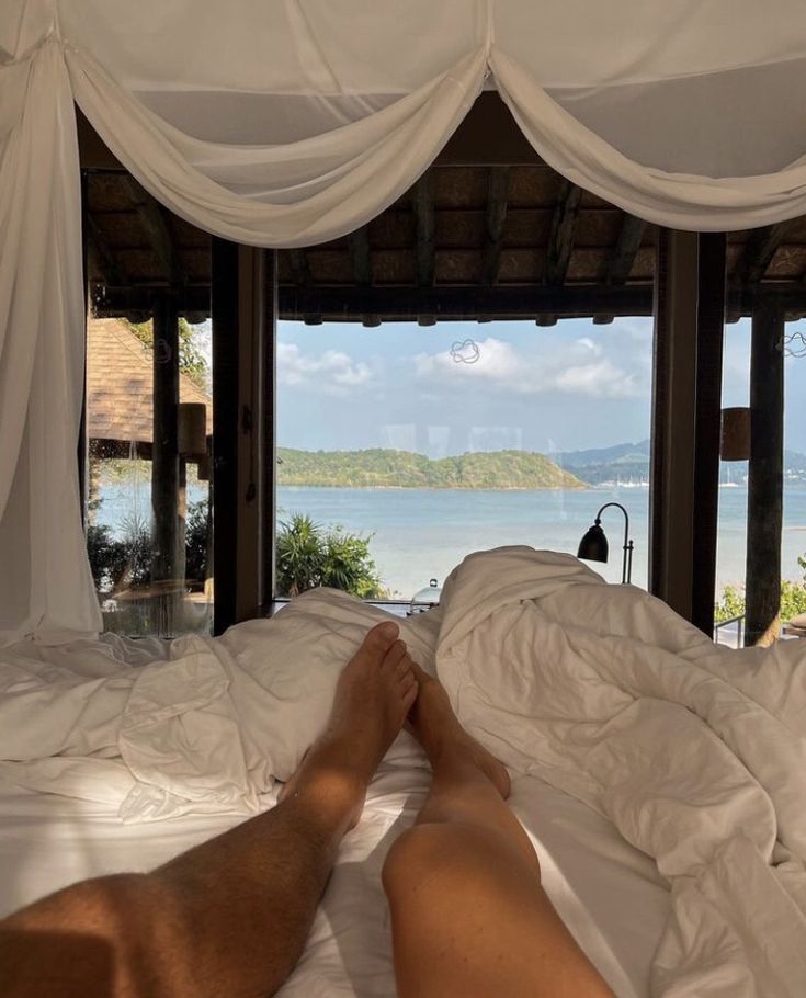 a man laying in bed with his feet up on the pillow and looking out at the ocean