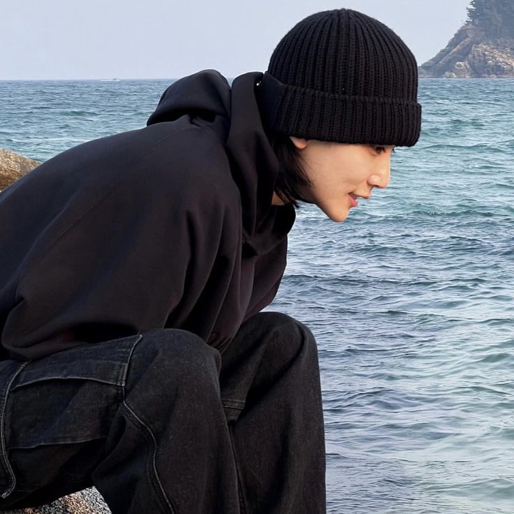 a young man sitting on top of a rock next to the ocean