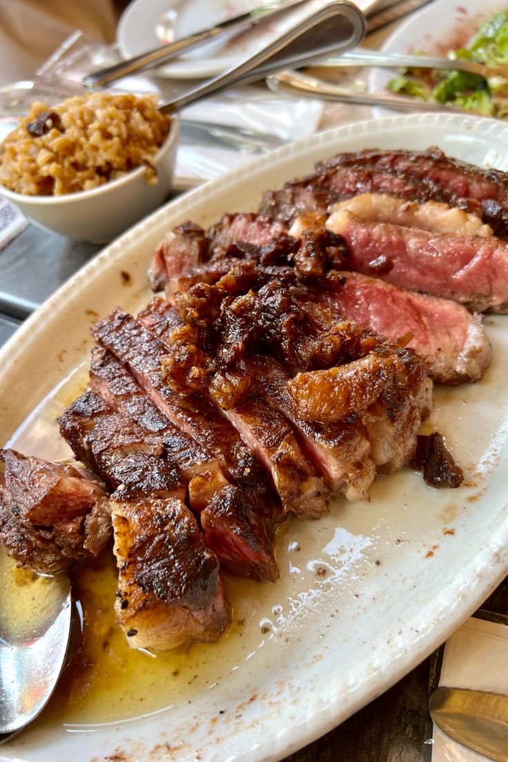 a white plate topped with meat next to a bowl of rice and spoons on top of a table