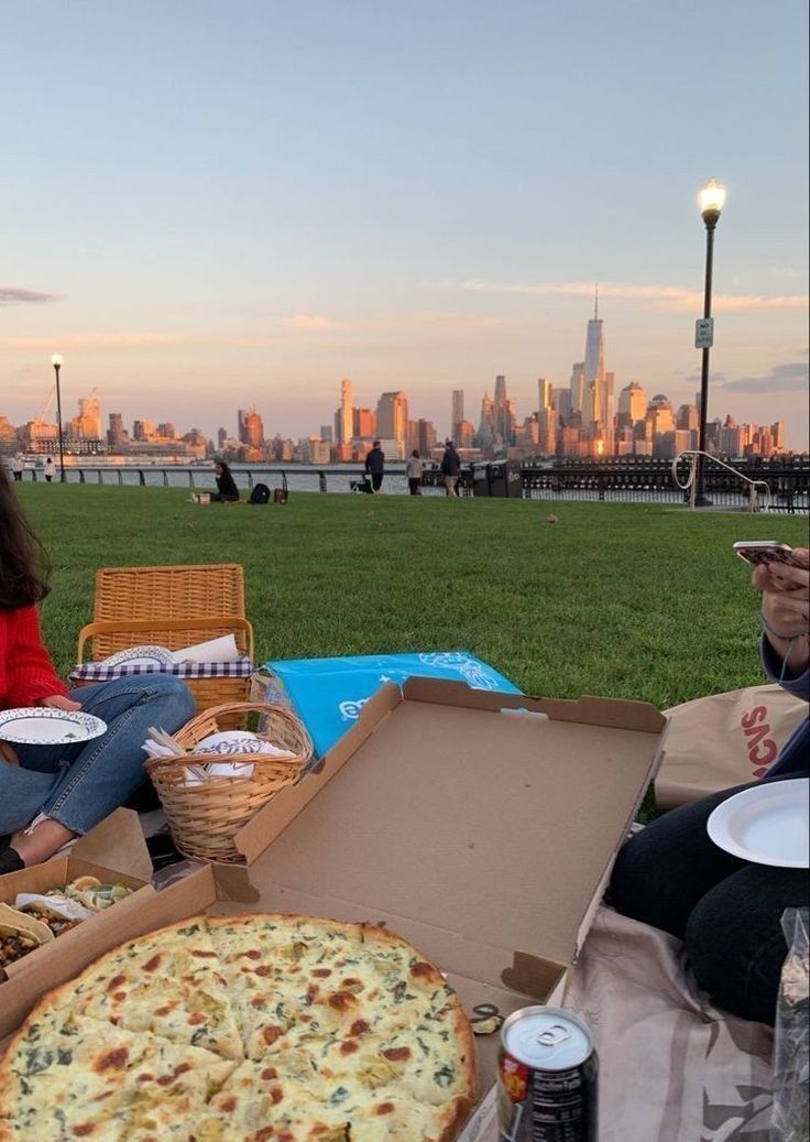 two people sitting on the grass eating pizza and drinking beer in front of a city skyline