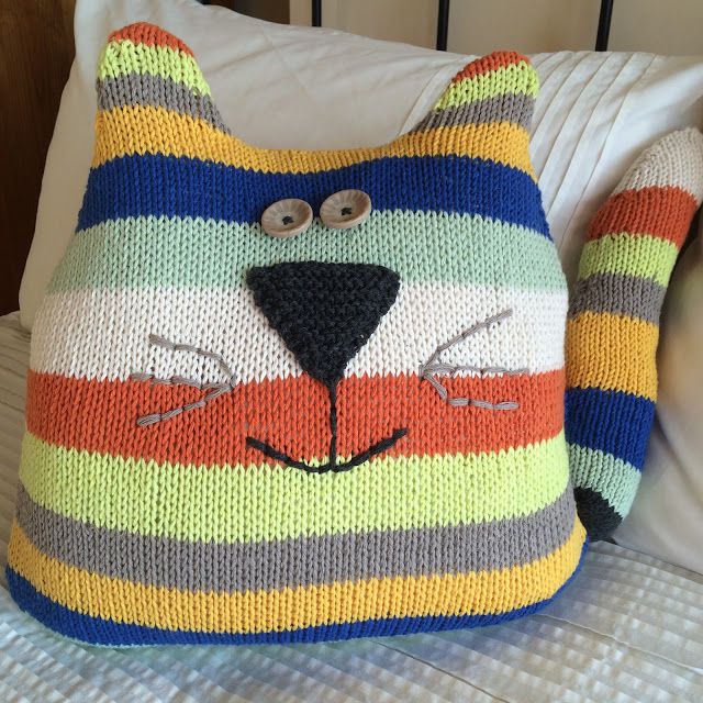 a colorful knitted cat pillow sitting on top of a bed next to two pillows