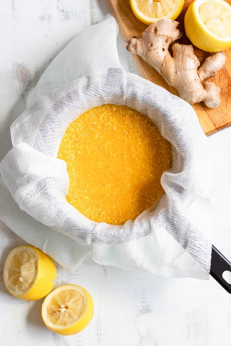 a bowl filled with yellow liquid next to sliced lemons and ginger on a cutting board