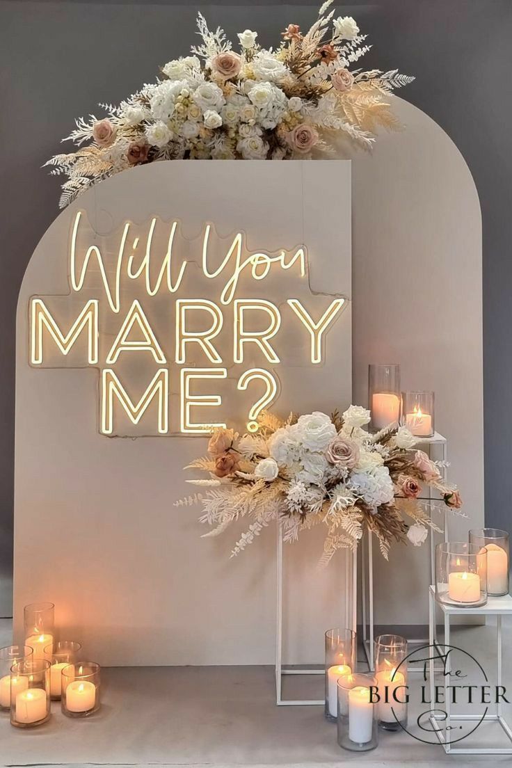 a lighted sign that says will you marry me? surrounded by candles and flowers in front of it