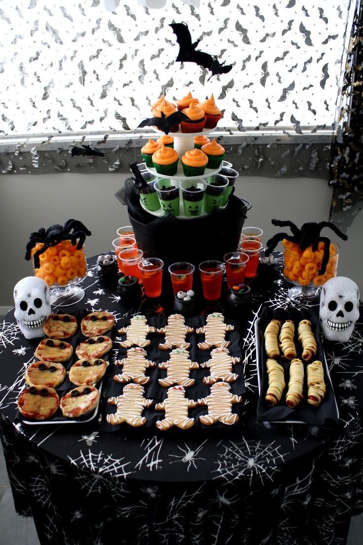 a table topped with halloween treats and desserts next to a window covered in spooky spider web