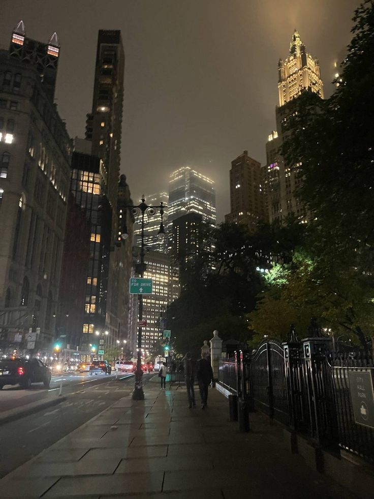 people are walking down the sidewalk in front of tall buildings at night time with lights on