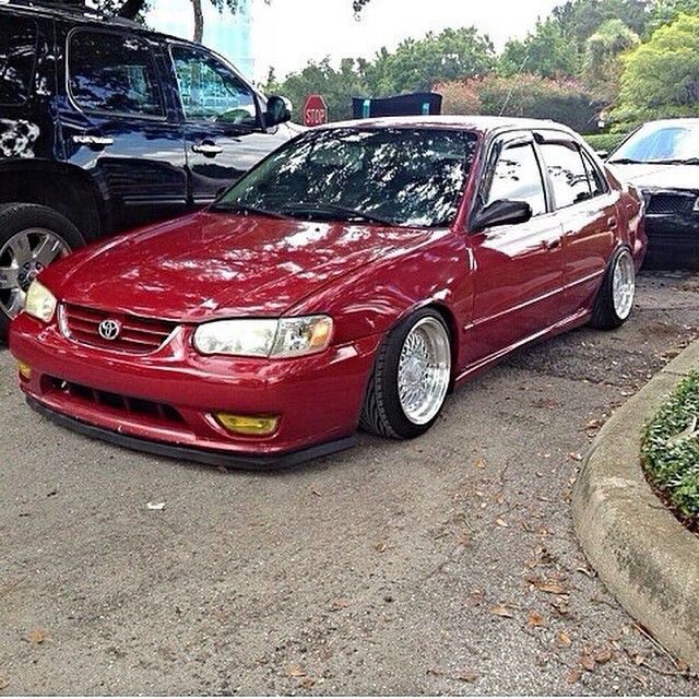 a red car parked next to other cars in a parking lot with trees and bushes