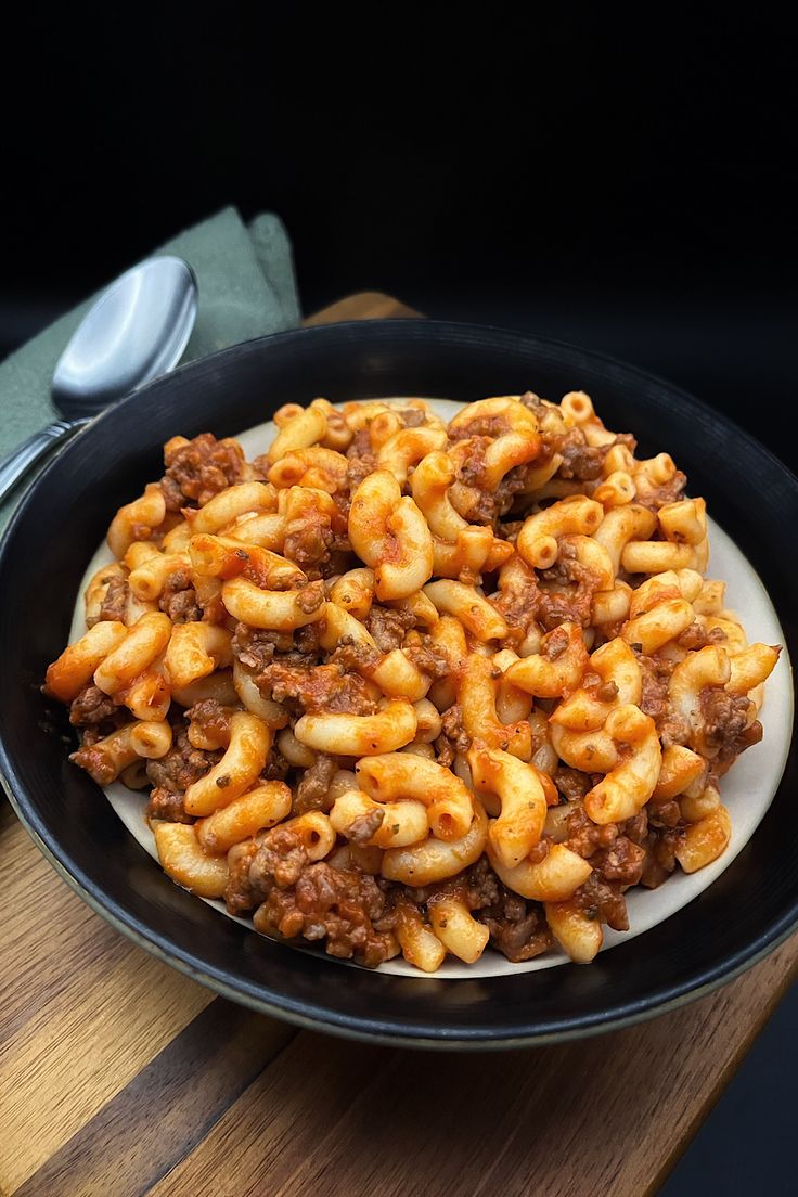 a black plate filled with pasta and meat on top of a wooden table next to a fork