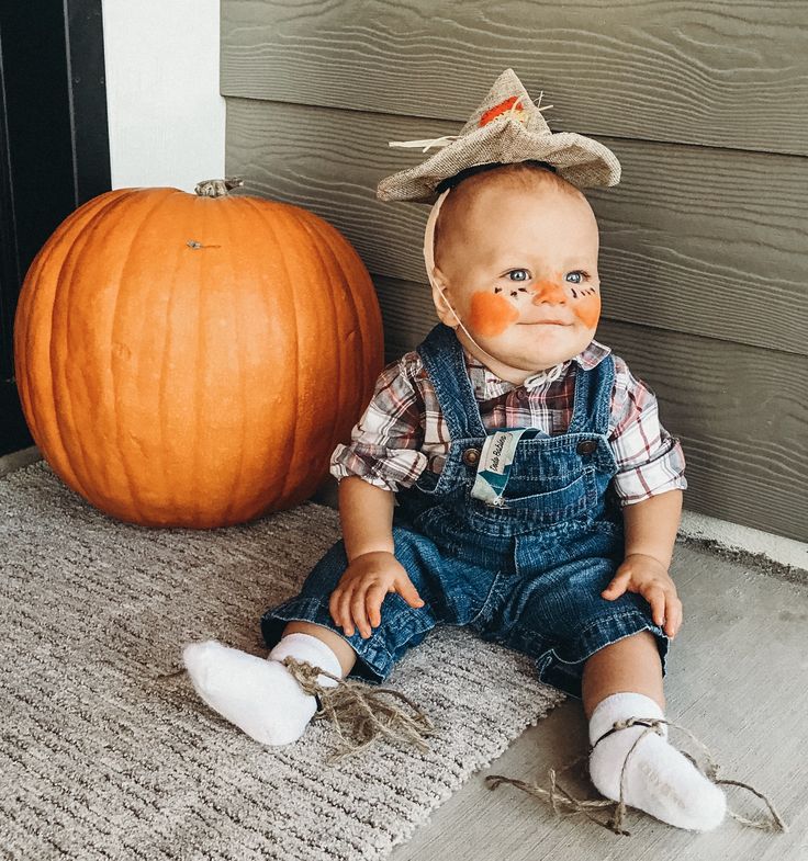 a baby sitting on the ground next to a pumpkin wearing a scarecrow's hat