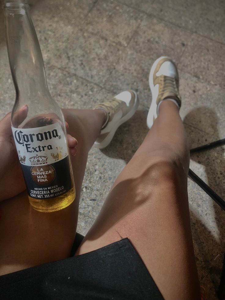 a person sitting on the ground with their legs crossed holding a bottle of corona beer