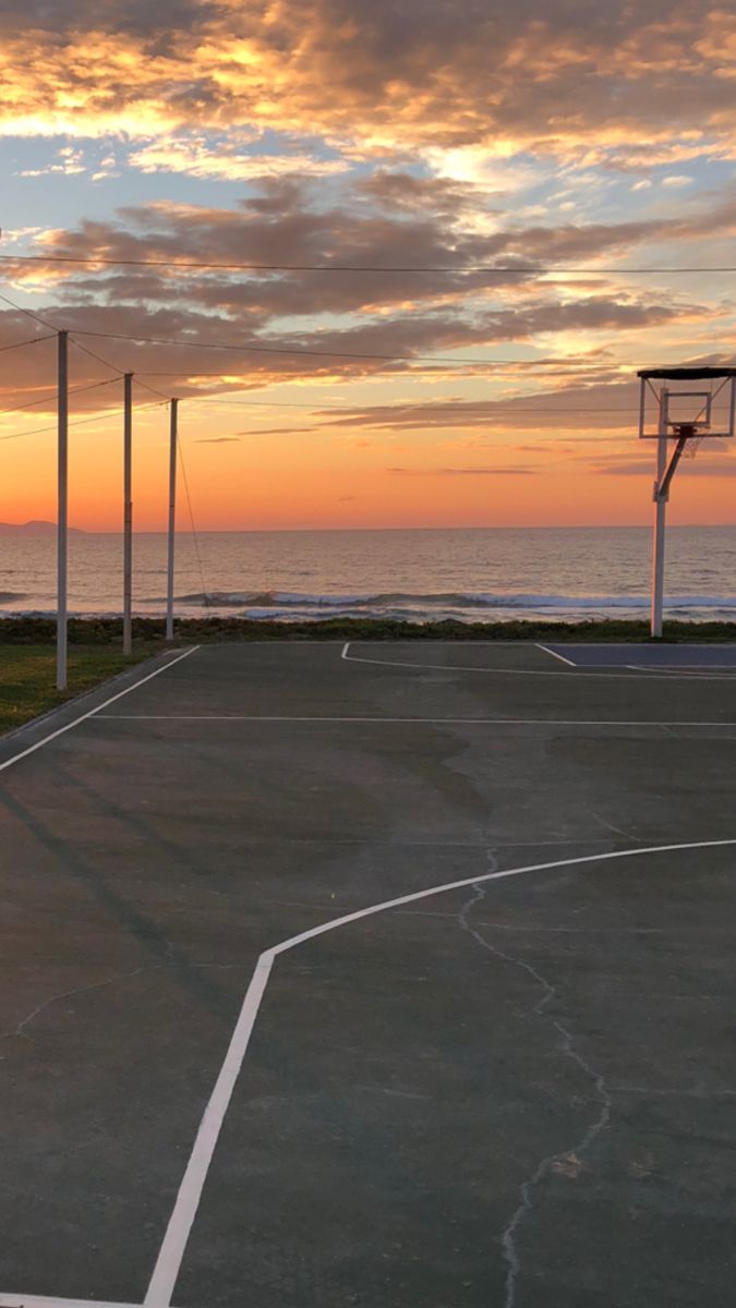 an empty basketball court with the sun setting in the background and clouds above it,