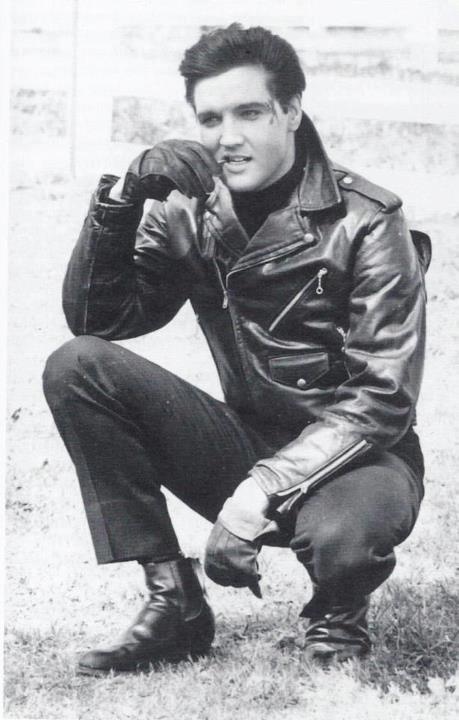 a black and white photo of a man in a leather jacket sitting on the ground