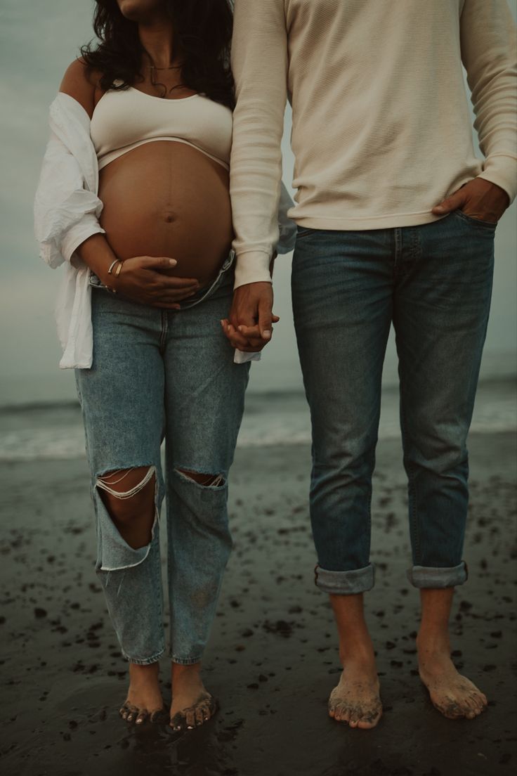 a pregnant couple holding hands while standing on the beach