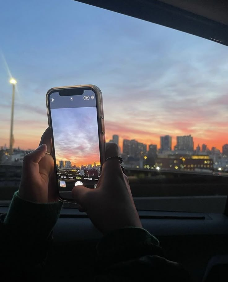 a person holding up a cell phone in front of a cityscape at sunset