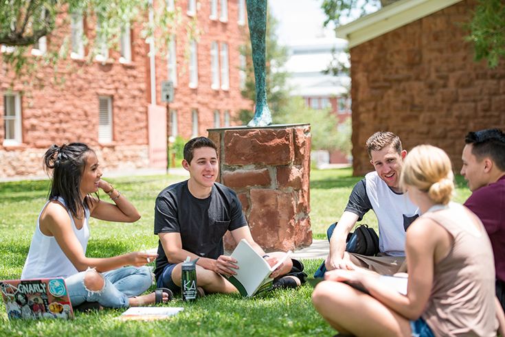 a group of young people sitting on the grass in front of a brick building talking to each other