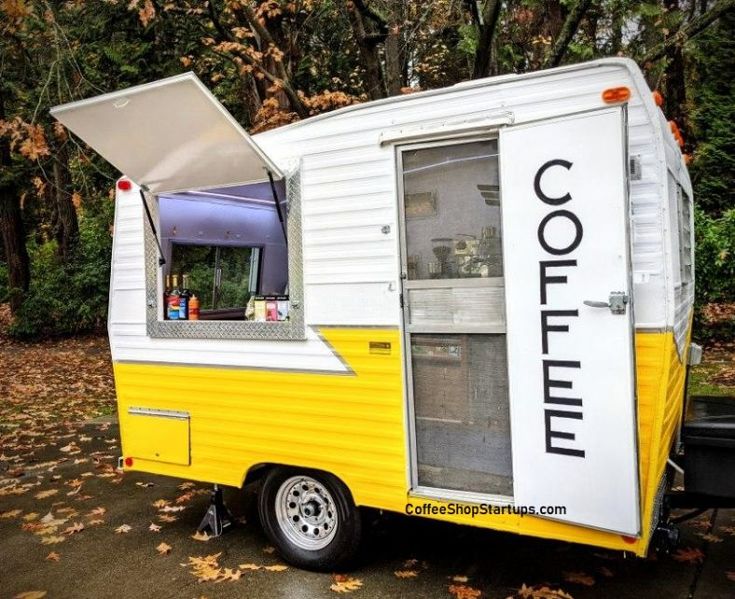 a small yellow and white food truck parked in front of trees with leaves on the ground