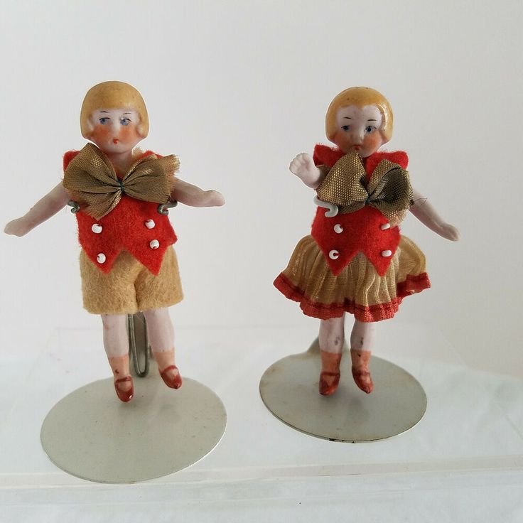 two figurines are standing on small bases, one is wearing a red dress and the other has a brown bow