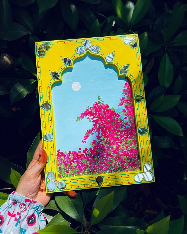 a person holding up a painting in front of some trees and bushes with pink flowers