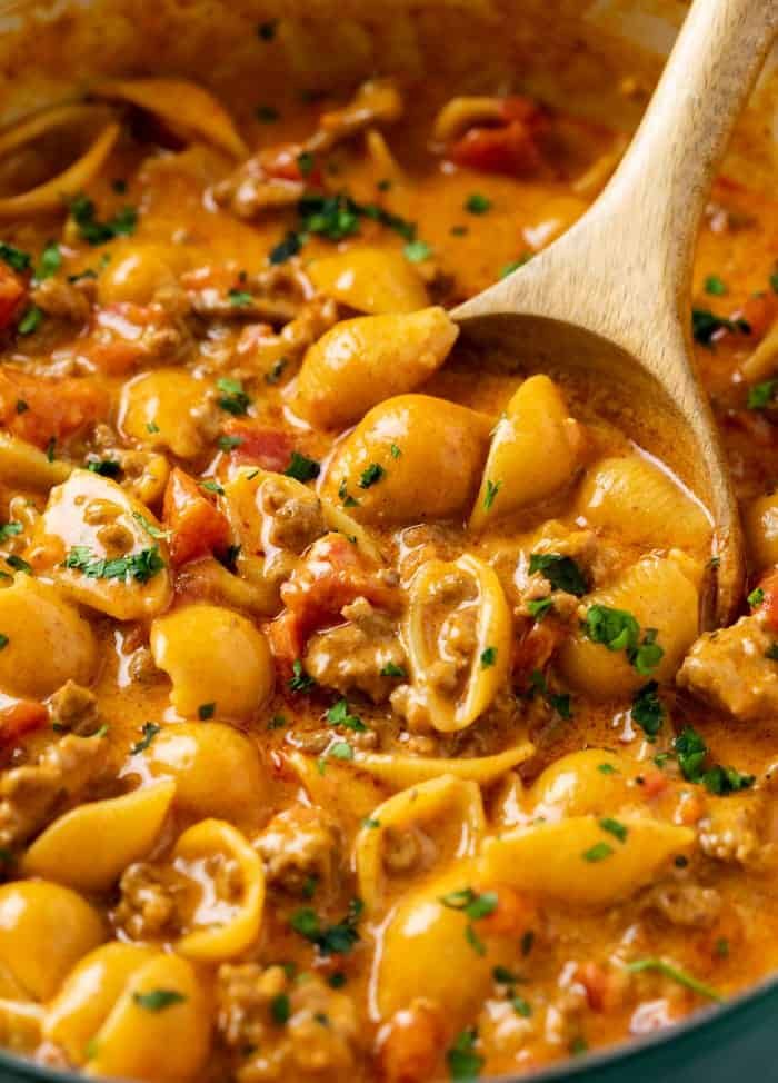 a wooden spoon in a pot filled with pasta and meat stew, topped with parsley