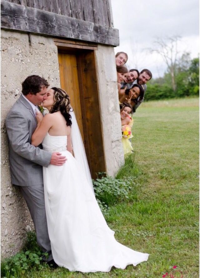 a bride and groom kissing in front of an outhouse with their wedding party behind them