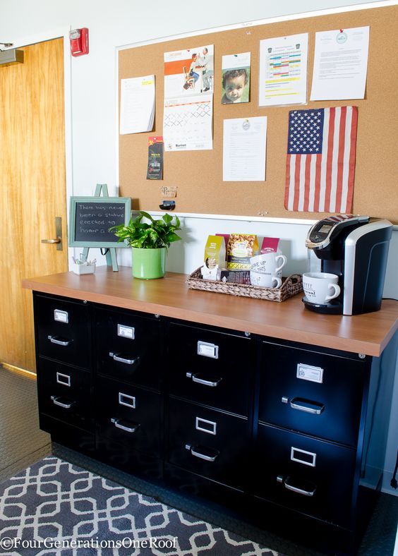 an office desk with several drawers and a coffee maker on top of it, in front of a corkboard bulletin board